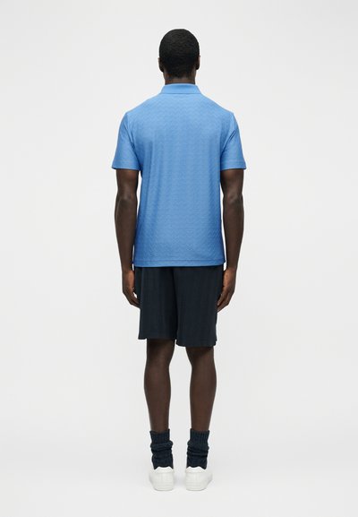 Άνδρας που φοράει ανοιχτό μπλε πόλο με υφή, μαύρα σορτς, σκούρες μαζεμένες κάλτσες και λευκά αθλητικά παπούτσια, στέκεται με την πλάτη γυρισμένη σε απλό φόντο.