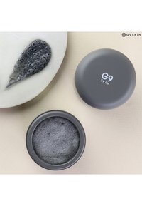 Conteneur cosmétique gris de forme ronde, étiqueté "G9 SKIN." Contenu : un produit noir texturé, présenté sur une surface blanche lisse.