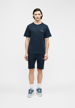 Νεαρός άνδρας όρθιος, φοράει μπλε ναυτικό t-shirt και σορτς με λευκές κάλτσες και ογκώδη αθλητικά σε απλό ανοιχτό φόντο.