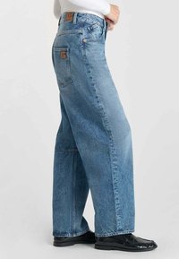 Jean large bleu clair en denim, avec une fermeture à boutons, cinq poches et un patch en cuir marron de la marque à l'arrière de la taille.