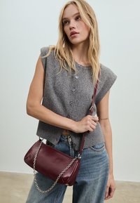 Szary dziergany bezrękawnik z przodu zapinany na guziki, połączony z luźnymi niebieskimi jeansami. Trzyma bordową skórzaną torebkę z łańcuszkowym paskiem.