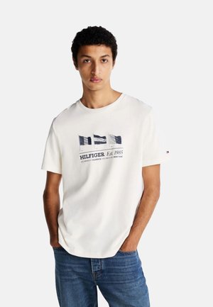 T-shirt bianca in cotone con stampa grafica di bandiere e testo "HILFIGER Est. 1985". Maniche corte, colletto rotondo e vestibilità rilassata.