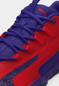 Κοντινό πλάνο ενός αθλητικού παπουτσιού σε κόκκινο και μωβ χρώμα με μωβ κορδόνια και κόκκινο λογότυπο Puma στη μωβ γλώσσα.