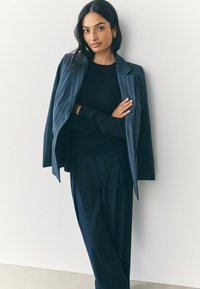 Marineblauw gestreepte blazer met notch-lapels, gedragen over een zwarte trui. Het model draagt op maat gemaakte donkere broeken, wat de gestructureerde silhouet benadrukt.