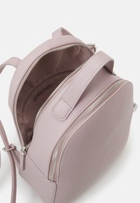 Mochila de cuero rosa claro con dos compartimentos con cremallera, uno parcialmente abierto que muestra el interior de tela marrón y un pequeño bolsillo interior con cremallera.