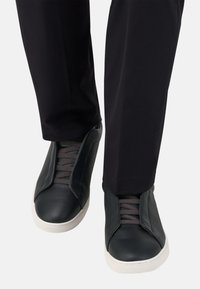 Baskets en cuir noir avec des lacets gris, présentant une texture lisse et une semelle en caoutchouc blanche. Portées avec un pantalon noir, mettant en avant un design élégant.