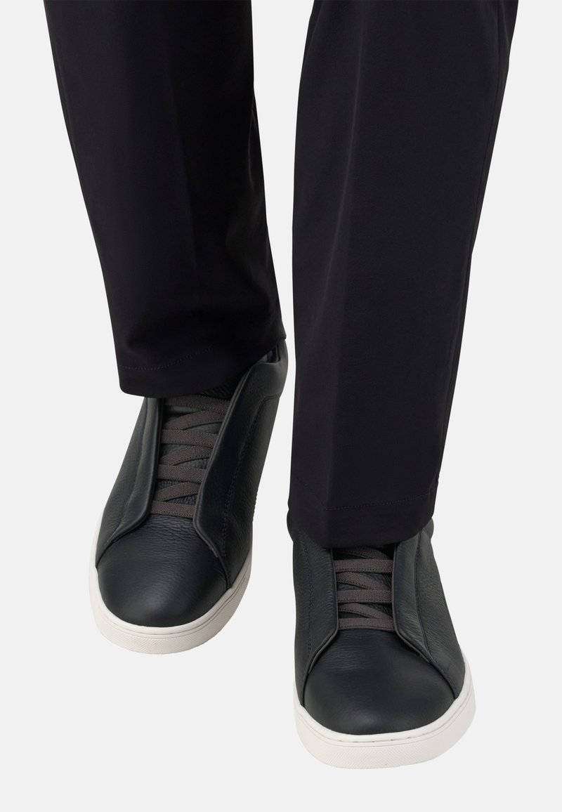 Baskets en cuir noir avec des lacets gris, présentant une texture lisse et une semelle en caoutchouc blanche. Portées avec un pantalon noir, mettant en avant un design élégant.