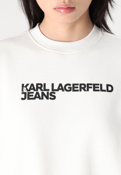 Бяла суичър с кръгло деколте, с черен текст "KARL LAGERFELD JEANS" в удебелен шрифт по предната част.