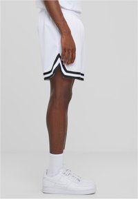 Shorts de sport en mesh blancs avec des rayures noires, dotés d'évents sur les côtés. Portés avec des baskets blanches et des chaussettes blanches arrivant aux chevilles.