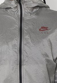 Nike Air bunda v stříbrno-černém mřížkovaném vzoru, s předním zipem a kapucí, s malým červeným logem na hrudi. Lehký materiál.