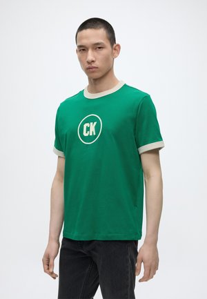Joven vestido con una camiseta verde de manga corta con ribetes blancos y un logo "CK", combinado con vaqueros negros, de pie frente a un fondo liso.