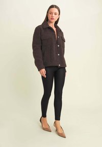 Chaqueta marrón de felpa con dos bolsillos en el pecho, cierres de botones y un collar. Llevada con leggings negros y tacones texturizados beige.