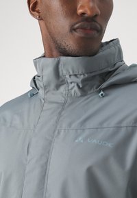 Vaude grå jakke med høy krage, teksturert stoff, justerbare snorstrammere og en liten logo på brystet.