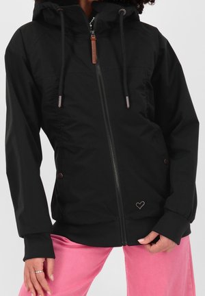 Frau trägt schwarze Kapuzenjacke mit Reißverschluss und Kordeln sowie pinke Hose, Hände locker an den Seiten, vor einfachem hellem Hintergrund.