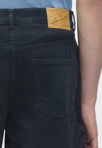 Πλάτη ατόμου που φοράει σκουρομπλε τζιν ντένιμ με δερμάτινη ετικέτα της μάρκας Les Deux στη μέση και ορατή πίσω τσέπη.
