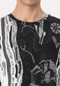 Svart og hvit strikket topp med abstrakte mønstre, varierte teksturer og rund hals. Viser en blanding av geometriske og organiske former.