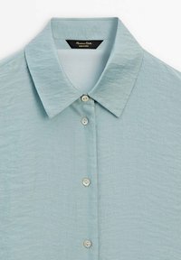 Chemise boutonnée bleu clair texturée avec col pointu et quatre boutons visibles sur le devant.