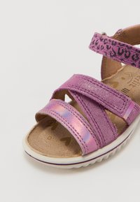 Lila sandaler med metallisk rosa accenter, med två breda remmar och en mjuk fotbädd. Sulan är vit med en vågig kant för bättre grepp.