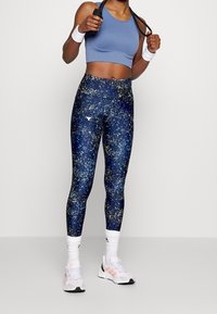 Atletische persoon in een blauwe crop top, gespikkelde marineblauwe leggings, witte Adidas sokken en witte sneakers, met een zwart springtouw in de hand.