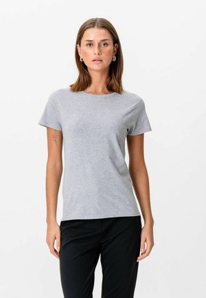 Kvinde med skulderlangt brunt hår iført en ensfarvet, lys grå kortærmet t-shirt og sorte bukser, stående foran en hvid baggrund.