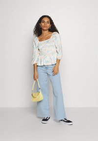 Mulher com cabelo encaracolado vestindo uma blusa floral com decote quadrado, jeans azul claro de perna larga, tênis pretos, segurando uma bolsa amarela com uma alça colorida.
