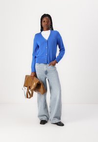Blue knitted cardigan with a v-neck, button closure, and ribbed hem, paired with light blue wide-leg jeans and a brown glossy tote bag.