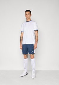 White athletic shirt with navy trim, paired with navy shorts. Worn with white socks displaying a pattern and black soccer cleats.