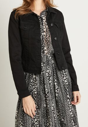 Kvinde iført en sort denimjakke over en sort og hvid blomstret kjole, stående med armene afslappet langs siderne.