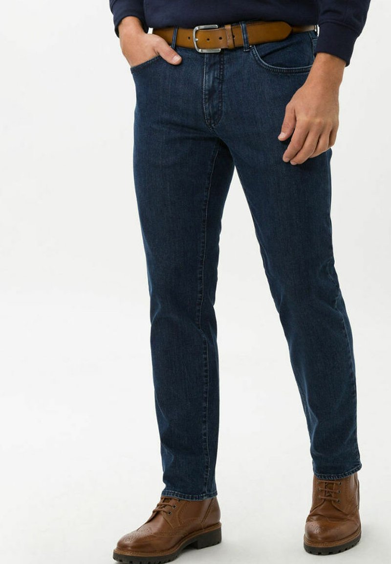 Jeans in denim blu scuro con taglio dritto, dotati di tasche frontali classiche e una cintura in pelle marrone. Stivali marroni stringati completano l'outfit.