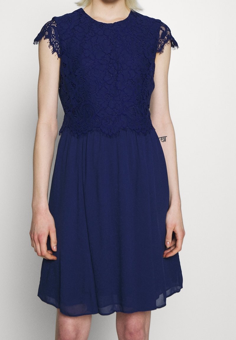 Robe bleu marine avec un haut en dentelle et des manches gigot, jupe en chiffon fluide, ourlet en dentelle festonnée et accents subtils de texture.