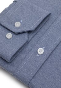 Niebieska koszula w geometryczny wzór z kołnierzykiem zapinanym na guziki, teksturalna tkanina oraz białe guziki przy mankietach i plisach. Szczegółowe przeszycia jako akcent.