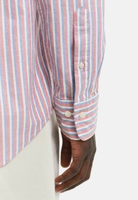 Chemise rayée avec des couleurs rose, bleu et blanc. Le tissu semble texturé avec des poignets à boutons. Détails de couture et de col visibles.