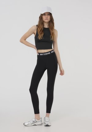 Top corto nero e leggings neri a vita alta con una cintura bianca "NON FERMARE ME", completati da scarpe da ginnastica argentate e un cappellino bianco.