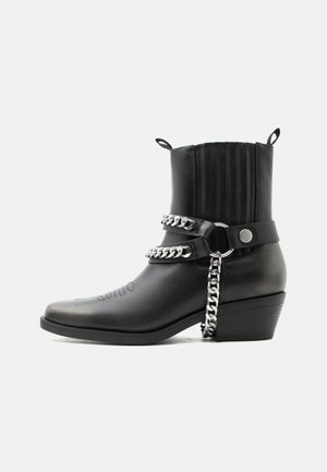 Bota de cuero negro para tobillo con tacón apilado, cadenas decorativas de plata, correas, tiradores y detalles cosidos en la punta y el caño.