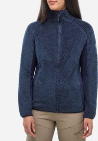 Femme portant une veste en polaire bleu marine zippée avec un col haut et un pantalon beige, se tenant devant un fond blanc uni.