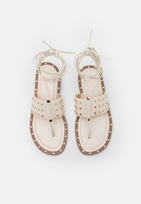 Sandali beige con due fasce larghe caratterizzate da borchie dorate e un puntale. Suola marrone con dettagli in borchie dorate lungo il bordo.