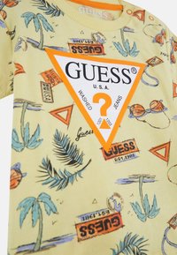 Chemise jaune à manches courtes avec des motifs tropicaux, des palmiers et des lunettes de soleil. Logo Guess orange en forme de triangle bien en évidence.