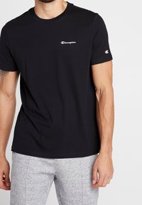 Man wearing black Champion t-shirt with logo on chest and sleeve, paired with light grey sweatpants, standing against white background.