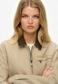 Veste beige avec fermeture éclair à l'avant, col en velours côtelé marron et patch sur la poche poitrine. Le modèle pose les bras croisés, mettant en valeur le design de la veste.