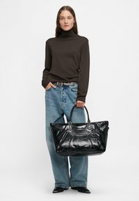 Frau trägt einen braunen Rollkragenpullover, locker sitzende blaue Jeans mit einem Nieten-Gürtel, schwarze Stiefel und hält eine große schwarze Lederhandtasche an ihrer Seite.