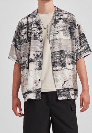 Man in een zwart-wit abstract patroon korte mouwen overhemd over een beige graphic t-shirt en zwarte cargo shorts.