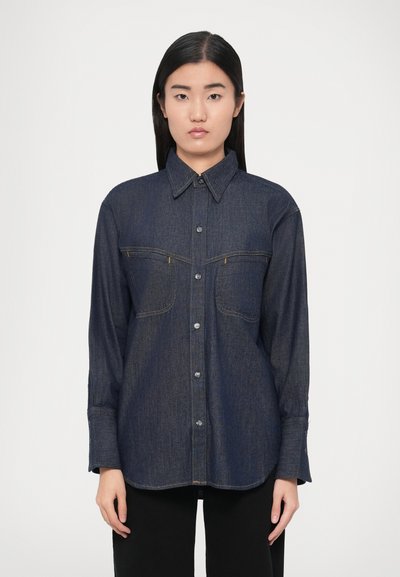 Camicia di jeans blu scuro con maniche lunghe, chiusura frontale con bottoni, colletto appuntito, due tasche sul petto e dettagli di cuciture sottili.