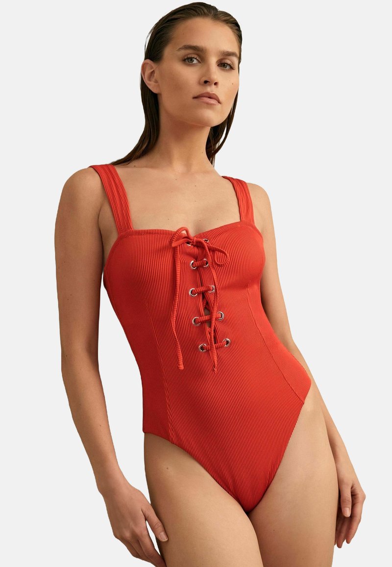 Le maillot de bain rouge côtelé présente une encolure carrée, des bretelles larges et un design à lacets à l'avant avec des œillets en métal. Les ouvertures de jambes hautes offrent une coupe élégante.