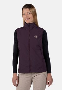 Violette ärmellose Weste mit hohem Kragen, Reißverschluss vorne und Logo. Getragen über einem langärmeligen schwarzen Shirt, das zwei seitliche Reißverschlusstaschen hat.