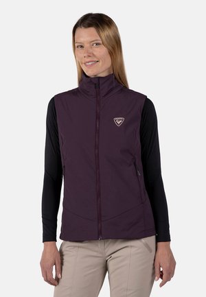 Violette ärmellose Weste mit hohem Kragen, Reißverschluss vorne und Logo. Getragen über einem langärmeligen schwarzen Shirt, das zwei seitliche Reißverschlusstaschen hat.