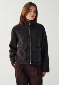 Giacca in pile grigia scura con colletto alto, zip frontale completa e due tasche anteriori. Tessuto morbido e strutturato con una vestibilità ampia.