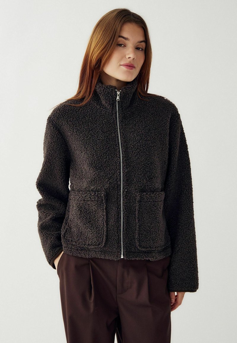 Giacca in pile grigia scura con colletto alto, zip frontale completa e due tasche anteriori. Tessuto morbido e strutturato con una vestibilità ampia.