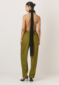 Moteris, apsidairanti atgal, vilkinti alyvuogių žalumos pritaikytas kelnes ir ilgą juodą šaliką, su užsirištu kaklu, atvira nugara ir juodais aukštakulniais.