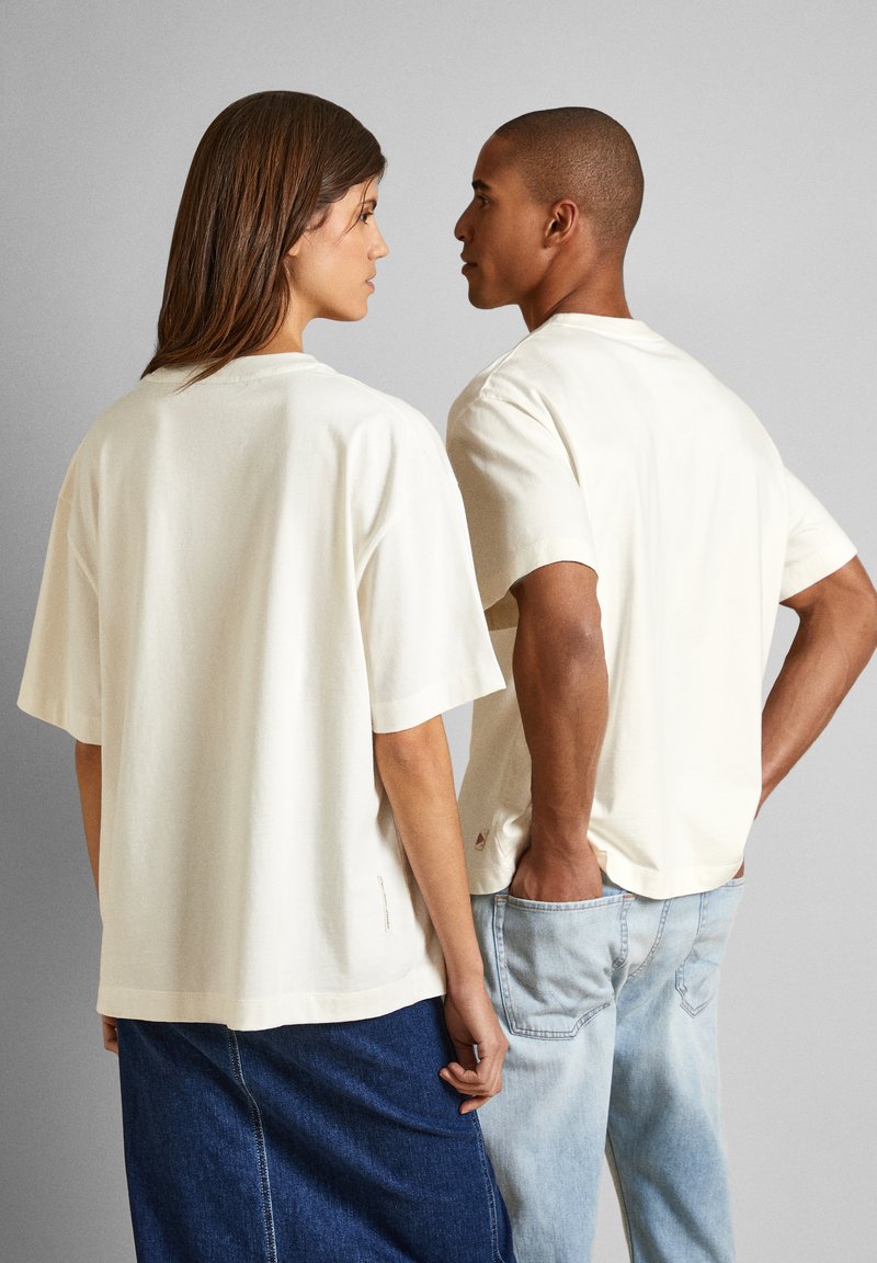 Helle beige oversized T-Shirts aus Baumwolle mit kurzen Ärmeln und lockerer Passform, von hinten mit Denim-Akzenten betrachtet.