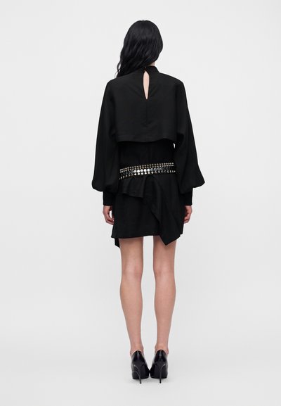 Mannequin vu de dos portant une mini robe drapée noire avec une ceinture ornée de bijoux et des talons hauts noirs.